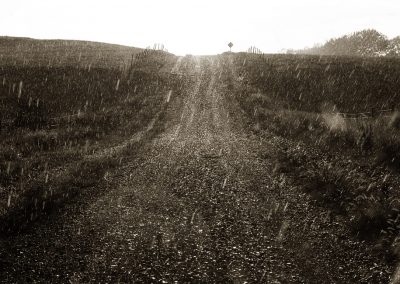 A view up a gravel road is textured by falling rain. A caution sign marks the top of the hill.